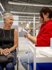 Oudere, lachende vrouw zit bij een vaccinatielocatie met de verpleegkundige naast zich.