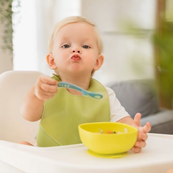 Blonde baby in kinderstoel met lepel in zijn handje en bakje eten op het tafelblad